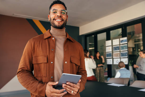 Happy young businessman looking at the camera while holding a digital tablet. Cheerful young businessman standing in a boardroom with his colleagues in the background.